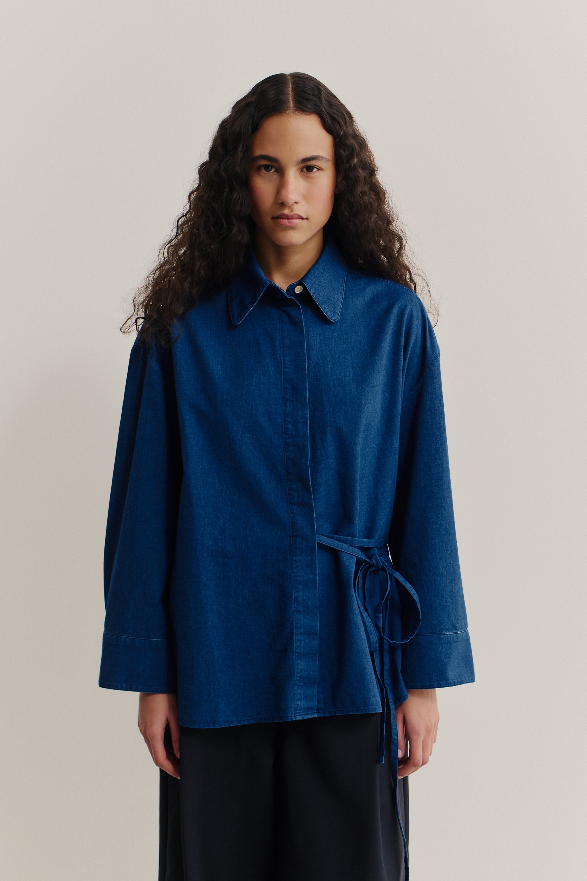 Women's long-sleeved shirt, washed indigo cotton, wide oversized fit, curved hem, and tie-waist detail.